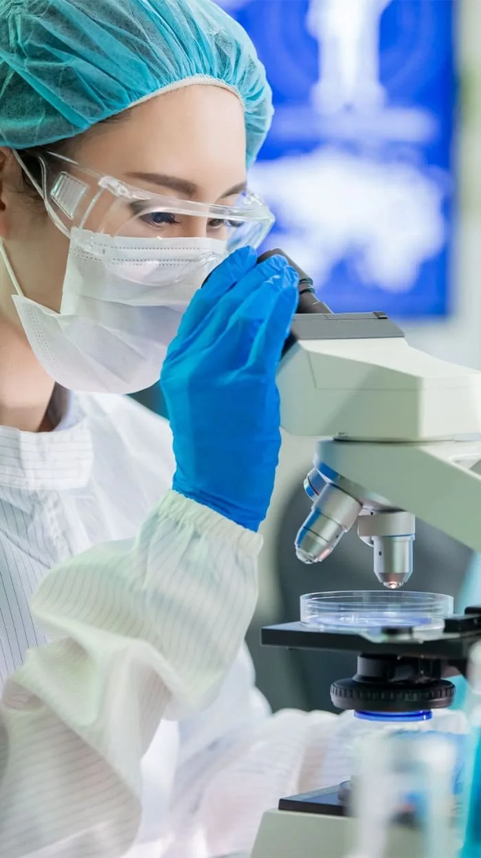 A laboratory scientist in protective equipment examining a sample through a microscope