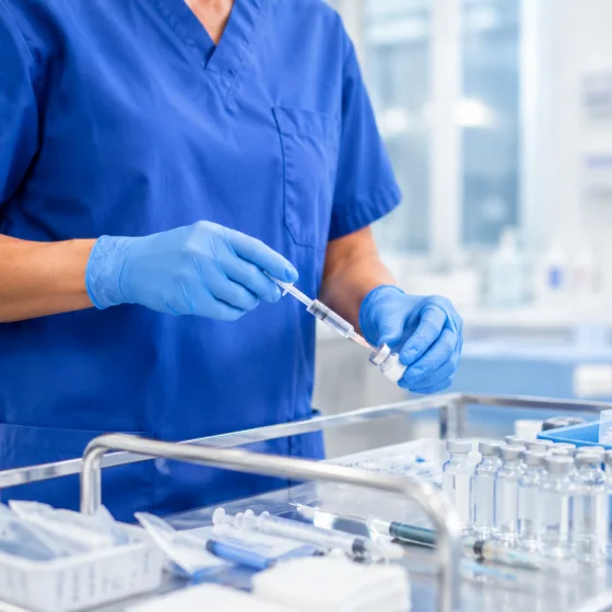 A nurse in scrubs and gloves preparing a syringe at a clinical supply trolley